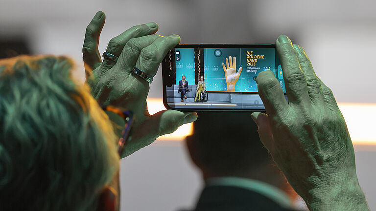 Nahaufnahme eines Zuschauers, der mit dem Smartphone eine Bühnenszene fotografiert. Auf dem Display sind die Moderatoren vor dem Logo „Die Goldene Hand 2025“ zu sehen. 