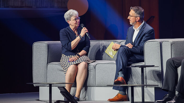 Eine Frau und ein Mann im Gespräch auf der Bühne. Beide sitzen auf grauen Sofas, die Frau hält ein Mikrofon, der Mann eine Programmkarte der Veranstaltung. 