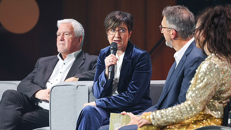 Vier Personen sitzen auf grauen Sofas auf der Bühne. Eine Frau im blauen Samtanzug spricht ins Mikrofon, während die anderen zuhören.