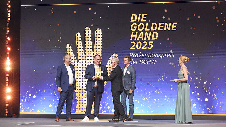 Übergabe der goldenen Hand-Trophäe auf der Bühne. Mehrere Personen stehen vor dem großen Veranstaltungslogo, während eine Moderatorin die Szene begleitet.