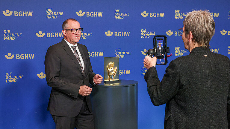 Ein Mann steht vor der blauen BGHW-Fotowand mit der goldenen Hand-Trophäe. Eine Frau filmt ihn mit dem Smartphone.