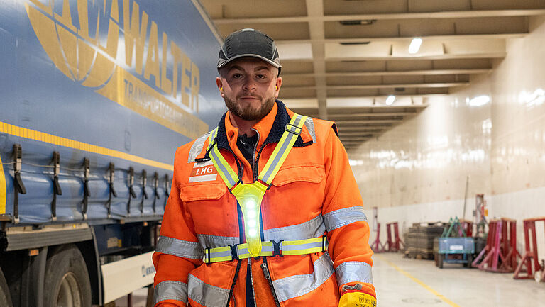Jastin Dos Santos Costa steht in Arbeitsmontur im hell erleuchteten Schiffsdeck. Über seiner Warnweste trägt er den Warnschutzgut mit LED-Leuchten – seine Idee für mehr Arbeitssicherheit auf dem Schiff. 