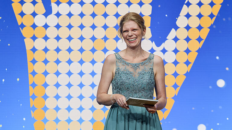 Moderatorin auf der Bühne der Preisverleihung „Die Goldene Hand 2025“ in türkisfarbenem Abendkleid, lächelnd vor dem Hintergrund mit dem goldenen Hand-Logo.