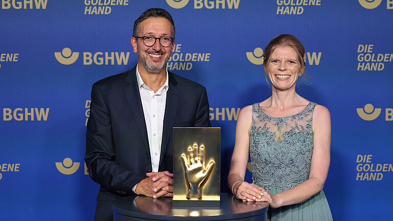 Ein Mann im Anzug und eine Frau im Abendkleid stehen gemeinsam vor einer BGHW-Fotowand; zwischen ihnen steht eine goldene Hand-Skulptur als Auszeichnung.
