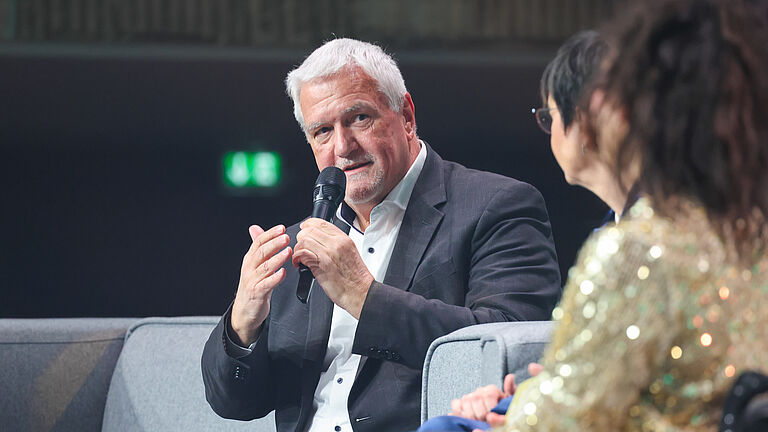 Ein Mann im dunklen Anzug spricht auf der Bühne ins Mikrofon und gestikuliert mit einer Hand. Neben ihm sitzen weitere Gesprächsteilnehmende.