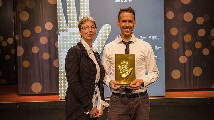 Zwei Personen stehen vor der BGHW Stellwand und halten den Goldene-Hand-Preis