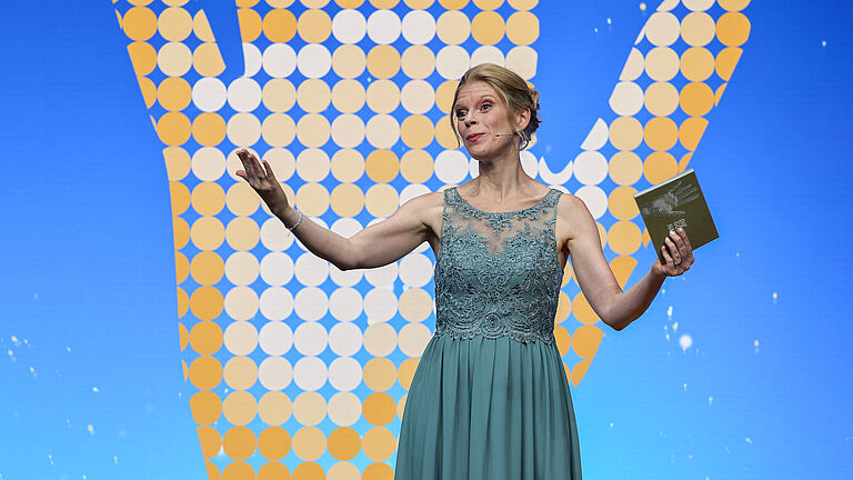 Moderatorin spricht auf der Bühne und gestikuliert mit den Händen, im Hintergrund das Logo „Die Goldene Hand 2025“ auf blauem Hintergrund.