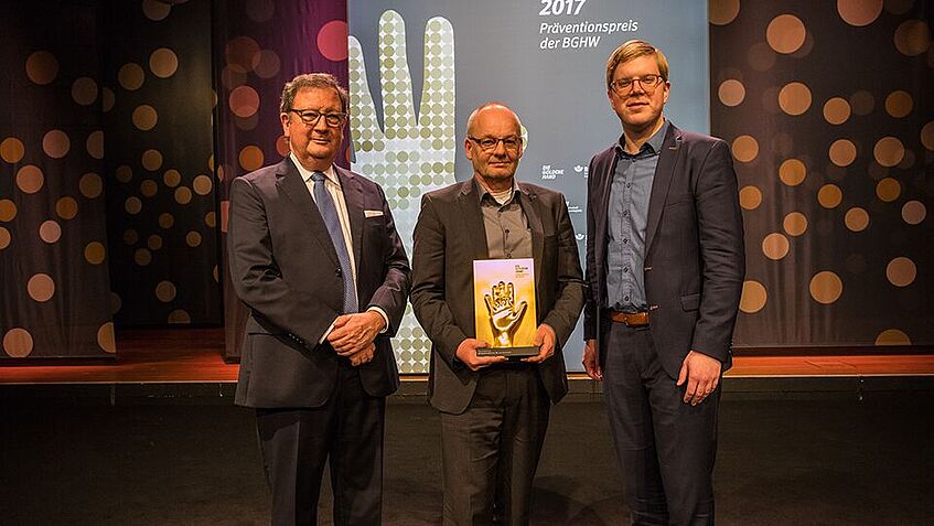 Drei Männer stehen vor der BGHW Stellwand und halten den Goldene-Hand-Preis