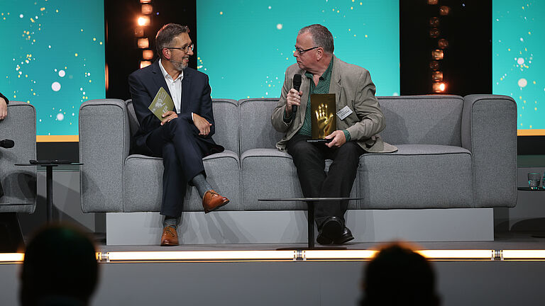 Ein Preisträger sitzt mit der goldenen Hand-Trophäe auf einem grauen Sofa und spricht ins Mikrofon. Neben ihm sitzt ein Moderator, der ihn lächelnd ansieht. Beide befinden sich auf einer beleuchteten Bühne im Rahmen der Preisverleihung.