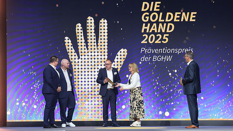 Preisverleihung auf der Bühne: Eine Frau überreicht einem Mann die Trophäe „Die Goldene Hand 2025“. Drei weitere Männer in Anzügen stehen daneben, während das große Logo der BGHW im Hintergrund leuchtet.