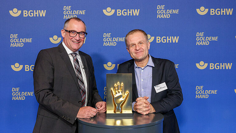 Zwei Männer posieren vor der BGHW-Fotowand mit der goldenen Hand-Trophäe und lächeln in die Kamera.