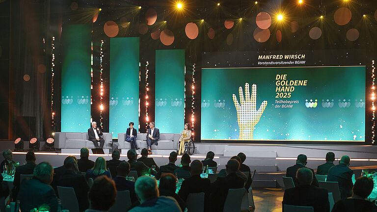 Podiumsdiskussion auf der Bühne der Veranstaltung „Die Goldene Hand 2025“. Vier Personen sitzen auf grauen Sofas, darunter eine Frau im Rollstuhl. Im Hintergrund leuchtet eine grün-goldene Bühnenwand mit dem Schriftzug „Manfred Wirsch – Vorstandsvorsitzender BGHW“ und „Teilhabepreis der BGHW“.