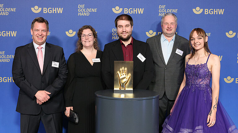 Fünf Personen stehen gemeinsam vor der BGHW-Fotowand und posieren mit der goldenen Hand-Trophäe.