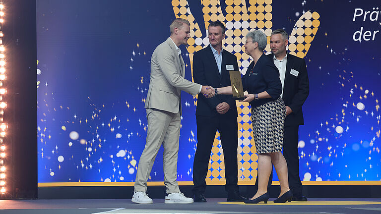 Podiumsgespräch auf der Bühne: Drei Männer und eine Moderatorin diskutieren über die ausgezeichneten Projekte. Einer der Männer hält die goldene Hand-Trophäe in den Händen. © BGHW, Michael Bauer