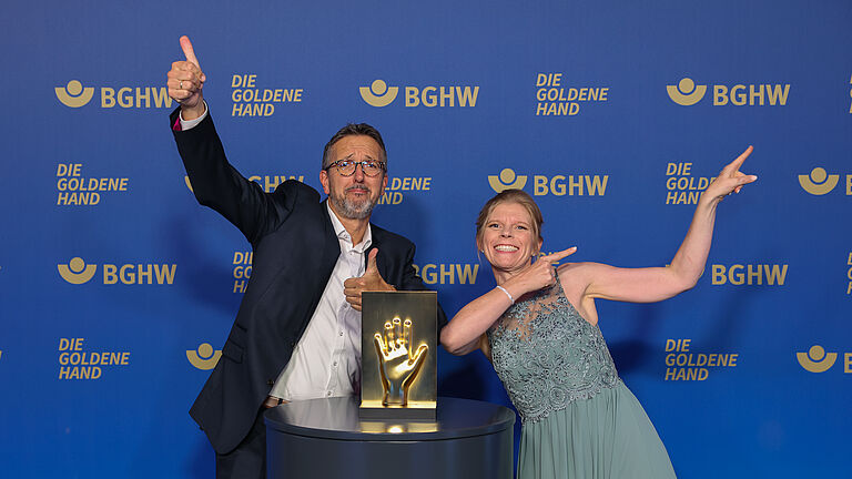 Ein Moderator und eine Moderatorin posieren gut gelaunt mit der goldenen Hand-Trophäe vor der offiziellen BGHW-Fotowand. Beide zeigen enthusiastische Posen und lächeln in die Kamera. © BGHW, Michael Bauer