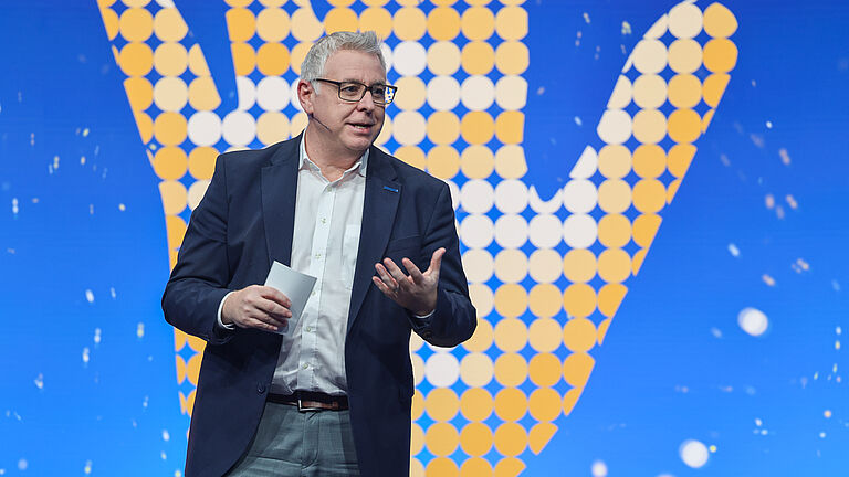 Ein Redner im Anzug spricht auf der Bühne der Preisverleihung „Die Goldene Hand 2025“. Im Hintergrund ist das große Logo der Veranstaltung in Form einer goldenen Hand auf blauem Grund zu sehen. © BGHW, Michael Bauer