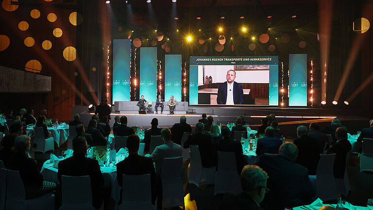 Bühnengespräch bei der Preisverleihung „Die Goldene Hand 2025“. Drei Personen sitzen auf Sofas und schauen auf eine große Leinwand, auf der ein Mann per Videozuschaltung erscheint. Über dem Bildschirm steht der Name „Johannes Koenen Transporte und Agrarservice, Rees“.