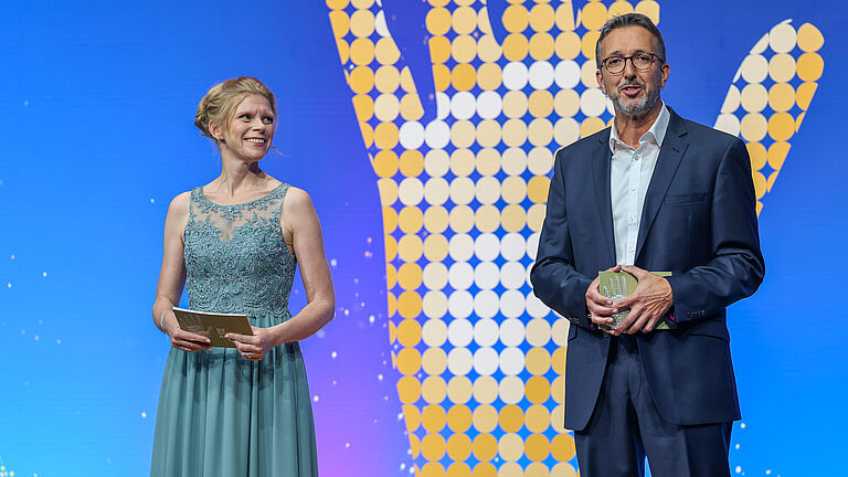Die beiden Moderatoren der Veranstaltung stehen nebeneinander auf der Bühne. Die Moderatorin trägt ein türkisfarbenes Abendkleid, der Moderator einen dunklen Anzug. Beide lächeln in Richtung Publikum, im Hintergrund das goldene Hand-Logo. © BGHW, Michael Bauer
