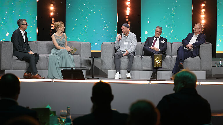 Vier Gesprächsteilnehmende sitzen auf der Bühne auf grauen Sofas. Ein Mann spricht ins Mikrofon, während die anderen aufmerksam zuhören. Die goldene Hand-Trophäe steht gut sichtbar vor der Gruppe.