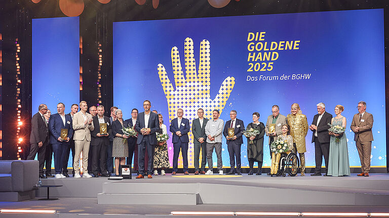 Abschlussfoto auf der Bühne: Alle Preisträgerinnen und Preisträger der „Goldenen Hand 2025“ stehen gemeinsam mit Moderatorinnen und Moderatoren vor dem großen Logo der Veranstaltung.