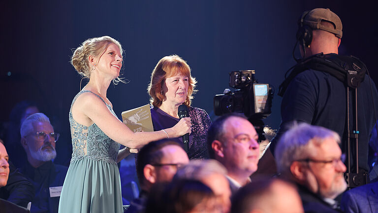 Die Moderatorin führt ein Interview mit einer Frau im Publikum, während eine Kamera das Gespräch aufzeichnet. Im Hintergrund sitzen weitere Gäste der Veranstaltung. 