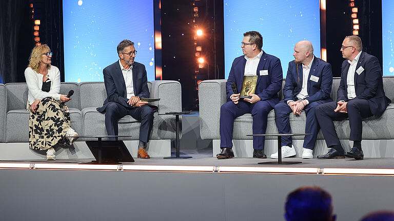 Podiumsgespräch auf der Bühne: Vier Männer und eine Frau diskutieren auf grauen Sofas. Zwei Männer halten die goldene Hand-Trophäe in den Händen. © BGHW, Michael Bauer