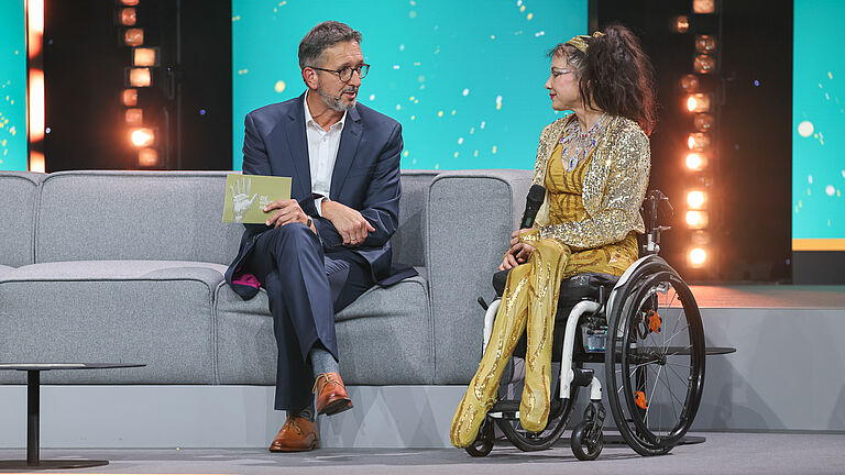 Ein Moderator spricht auf der Bühne mit einer Frau im goldenen Outfit, die im Rollstuhl sitzt. Beide wirken konzentriert und sind im Gespräch vertieft. 