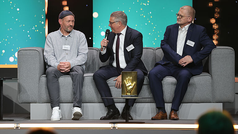 Drei Männer sitzen auf grauen Sofas auf der Bühne, einer spricht ins Mikrofon. Vor ihnen steht die goldene Hand-Trophäe auf einem kleinen Tisch.