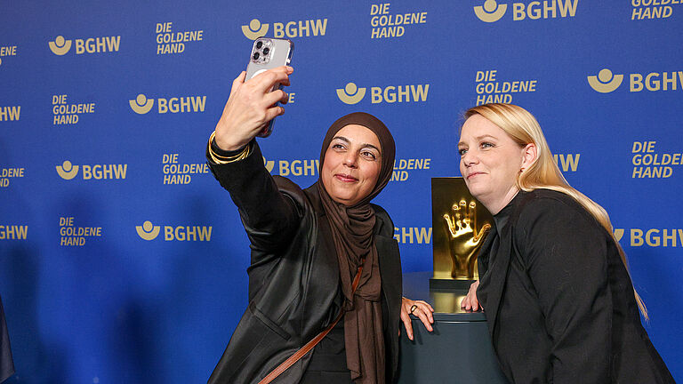 Zwei Frauen machen ein Selfie vor der blauen BGHW-Fotowand mit der goldenen Hand-Trophäe im Hintergrund.