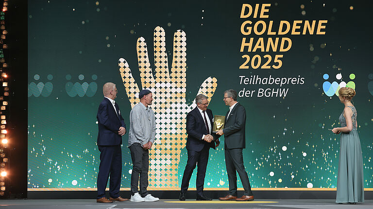 Auf der Bühne wird die goldene Hand-Trophäe überreicht. Mehrere Personen stehen nebeneinander, während ein Mann einem anderen gratuliert und die Trophäe überreicht.