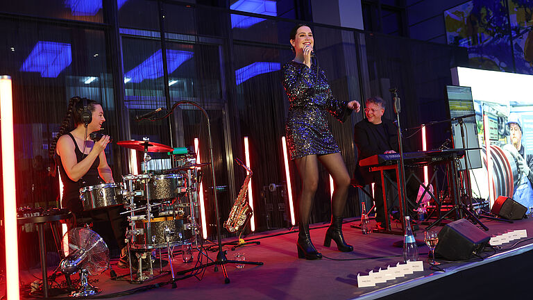 Die gleiche Live-Band spielt auf der Bühne. Die Schlagzeugerin lacht, während die Sängerin singt. Der Keyboarder begleitet sie. Hinter ihnen sorgen rote Lichtstäbe für eine moderne Bühnenatmosphäre.