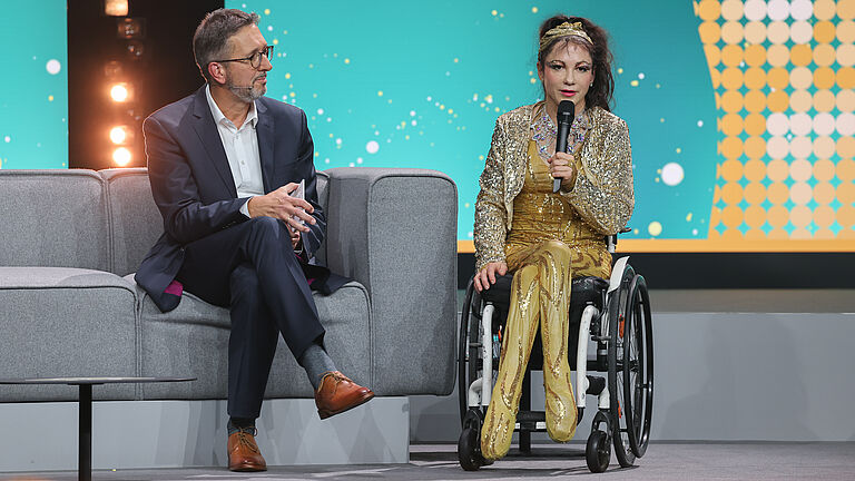Eine Frau im Rollstuhl und ein Mann im Anzug sitzen auf der Bühne und sprechen ins Mikrofon. Die Frau trägt ein goldenes, glitzerndes Outfit.