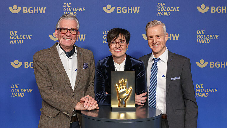 Drei Personen stehen vor einer blauen Fotowand mit dem Logo „Die Goldene Hand“. Sie lächeln in die Kamera und präsentieren die goldene Hand-Trophäe auf einem Podest. 