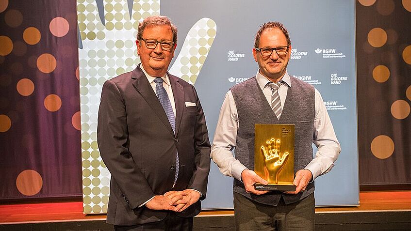 Zwei Männer stehen vor der BGHW Stellwand und halten den Goldene-Hand-Preis