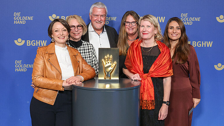 Eine Gruppe aus sechs Personen steht vor der BGHW-Fotowand und präsentiert die goldene Hand-Trophäe auf einem Podest.