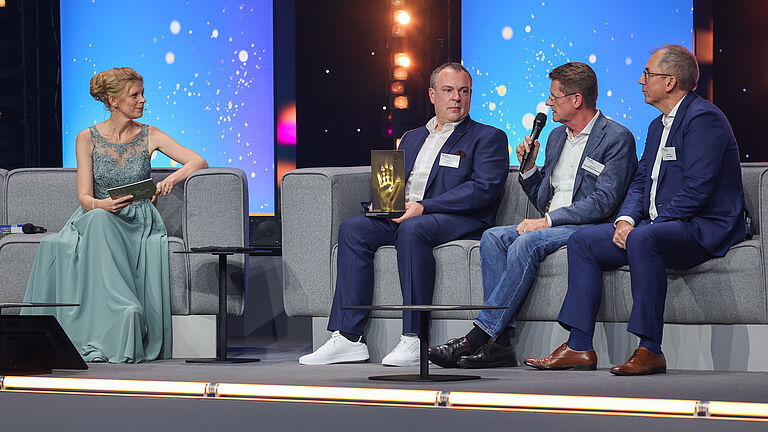 Ein Mann im hellen Anzug schüttelt auf der Bühne einer Frau die Hand, während er die goldene Hand-Trophäe entgegennimmt. Zwei weitere Männer im Hintergrund beobachten die Szene. 