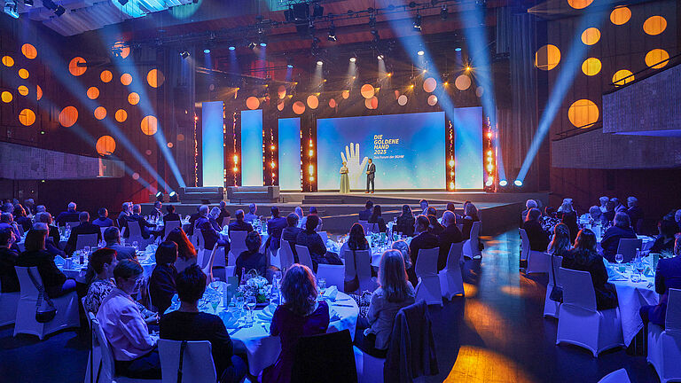 Blick in den Veranstaltungssaal der Preisverleihung „Die Goldene Hand 2025“. Das Publikum sitzt an festlich gedeckten Tischen, während die Moderatoren auf der Bühne stehen.