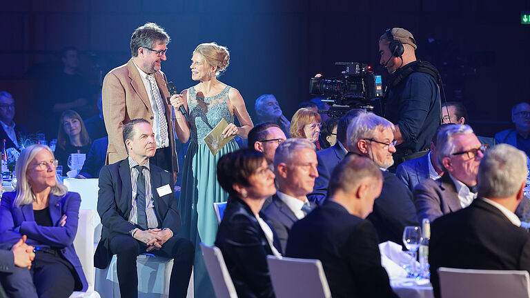 Die Moderatorin interviewt einen Mann im hellbraunen Sakko mitten im Publikum. Viele Gäste schauen gespannt zu, während Kameras die Szene aufnehmen. 
