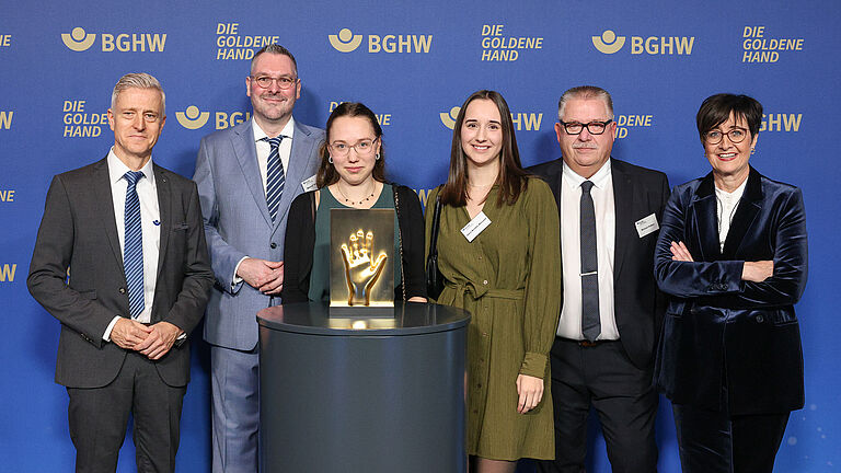 Sechs Personen posieren gemeinsam vor einer blauen Fotowand mit dem BGHW-Logo und dem Schriftzug „Die Goldene Hand“. In der Mitte steht die goldene Hand-Trophäe auf einem Podest. 