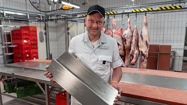 Mitarbeiter steht in einem Fleischbetrieb und posiert mit einem Schutzblech in der Hand