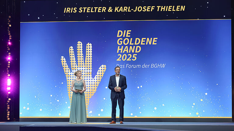Zwei Moderatoren stehen auf der Bühne der Preisverleihung „Die Goldene Hand 2025“. Auf der Leinwand hinter ihnen sind ihre Namen „Iris Stelter & Karl-Josef Thielen“ sowie das Logo der Veranstaltung mit der goldenen Hand zu sehen.