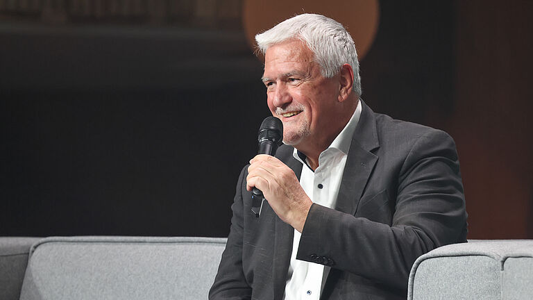 Ein älterer Mann im dunklen Anzug spricht lächelnd ins Mikrofon, während er auf einem grauen Sofa auf der Bühne sitzt.