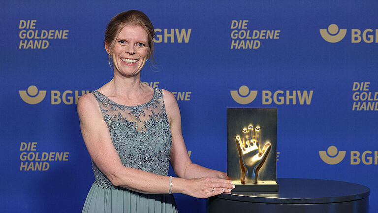 Frau in einem hellblauen Abendkleid posiert vor einer blauen Fotowand mit dem Logo ‚Die Goldene Hand‘ und der BGHW; neben ihr steht eine goldfarbene Award-Skulptur in Form einer Hand.
