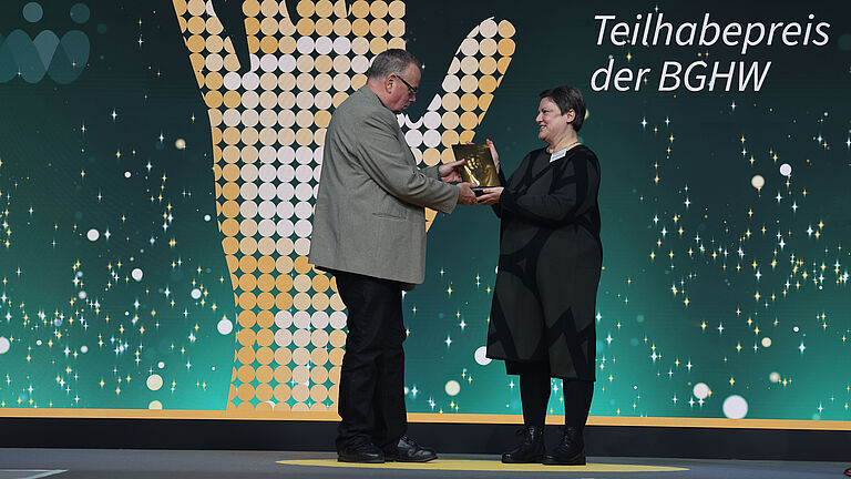 Eine Frau in schwarzem Outfit nimmt auf der Bühne die goldene Hand-Trophäe entgegen. Ein Mann im grauen Sakko überreicht ihr die Auszeichnung. Im Hintergrund ist eine große, stilisierte Handgrafik sowie der Text „Teilhabepreis der BGHW“ zu sehen.