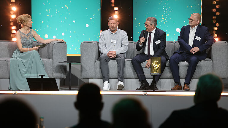 Eine Moderatorin im türkisfarbenen Abendkleid sitzt auf der Bühne und spricht mit drei Männern. Einer von ihnen hält ein Mikrofon, die goldene Hand-Trophäe steht vor ihnen.