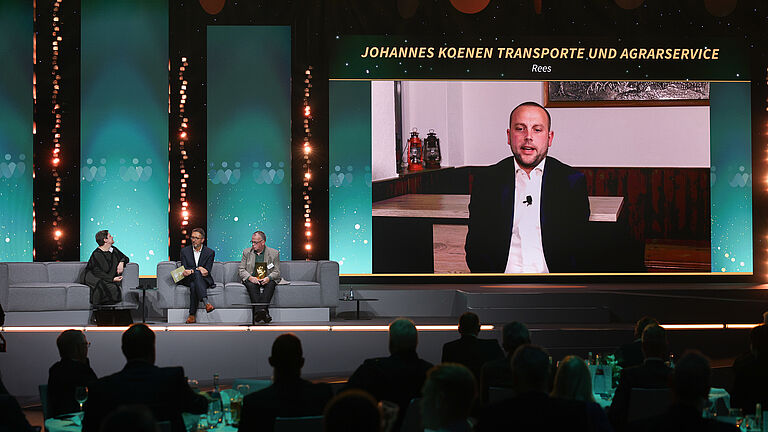 Bühnengespräch bei der Preisverleihung „Die Goldene Hand 2025“. Drei Personen sitzen auf Sofas und schauen auf eine große Leinwand, auf der ein Mann per Videozuschaltung erscheint. Über dem Bildschirm steht der Name „Johannes Koenen Transporte und Agrarservice, Rees“.