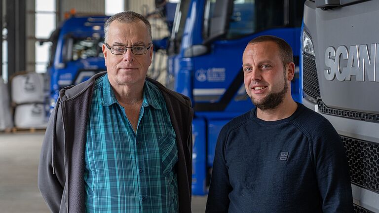 Zwei Männer stehen nebeneinander in einer Fahrzeughalle und schauen in die Kamera. Im Hintergrund stehen parkende Lkw. 