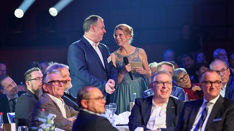 Publikumsszene: Eine Moderatorin im türkisfarbenen Kleid spricht mit einem Mann im Anzug, der im Publikum steht. Die Gäste am Tisch lachen und applaudieren. © BGHW, Michael Bauer